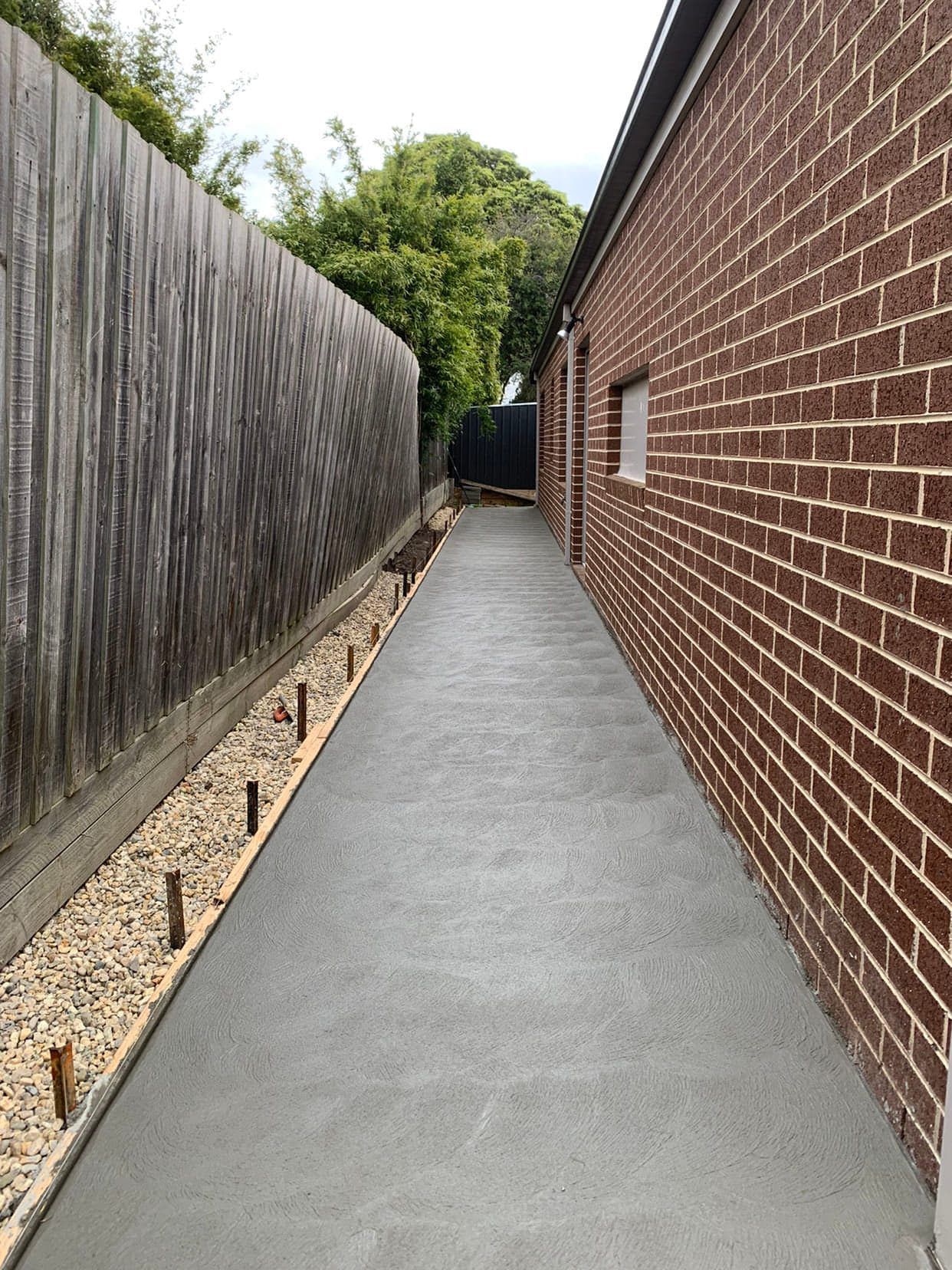 Long, gray concrete pathway between a brick building and a wooden fence, with gravel and wooden stakes along the fence.
