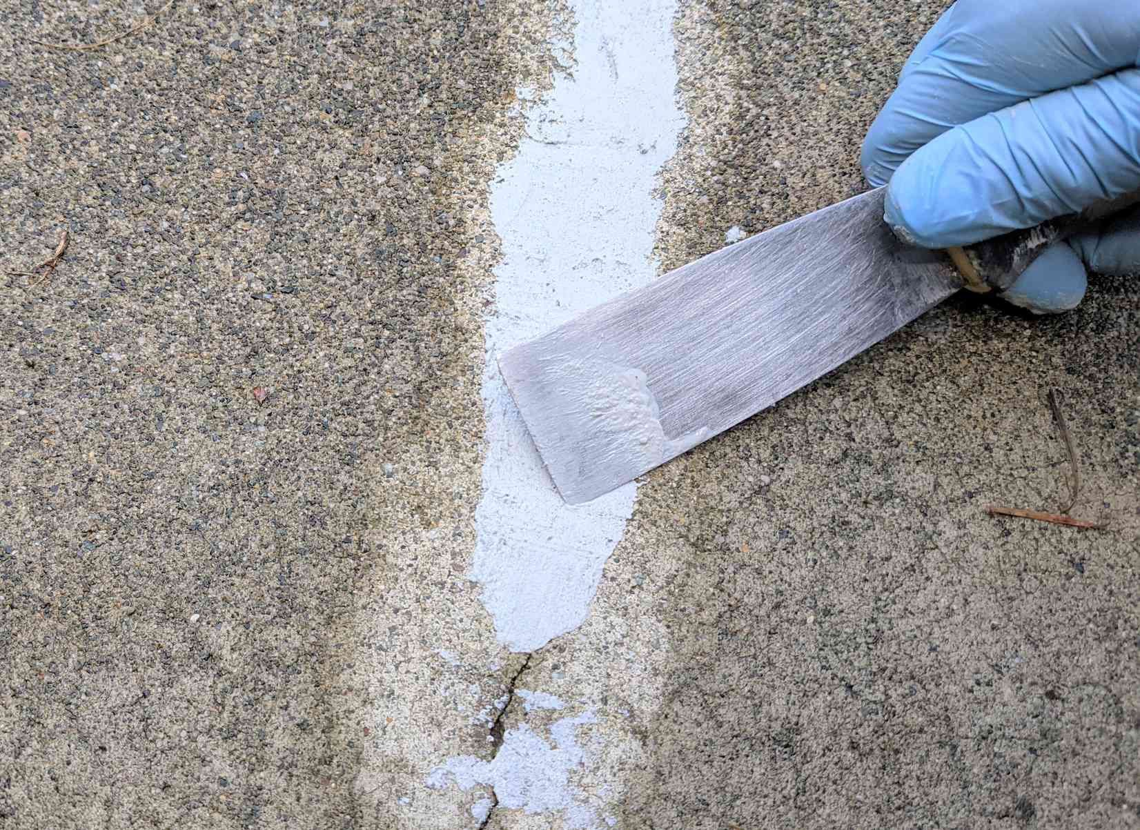 Gloved hand using a putty knife to apply white sealant to a crack in concrete.