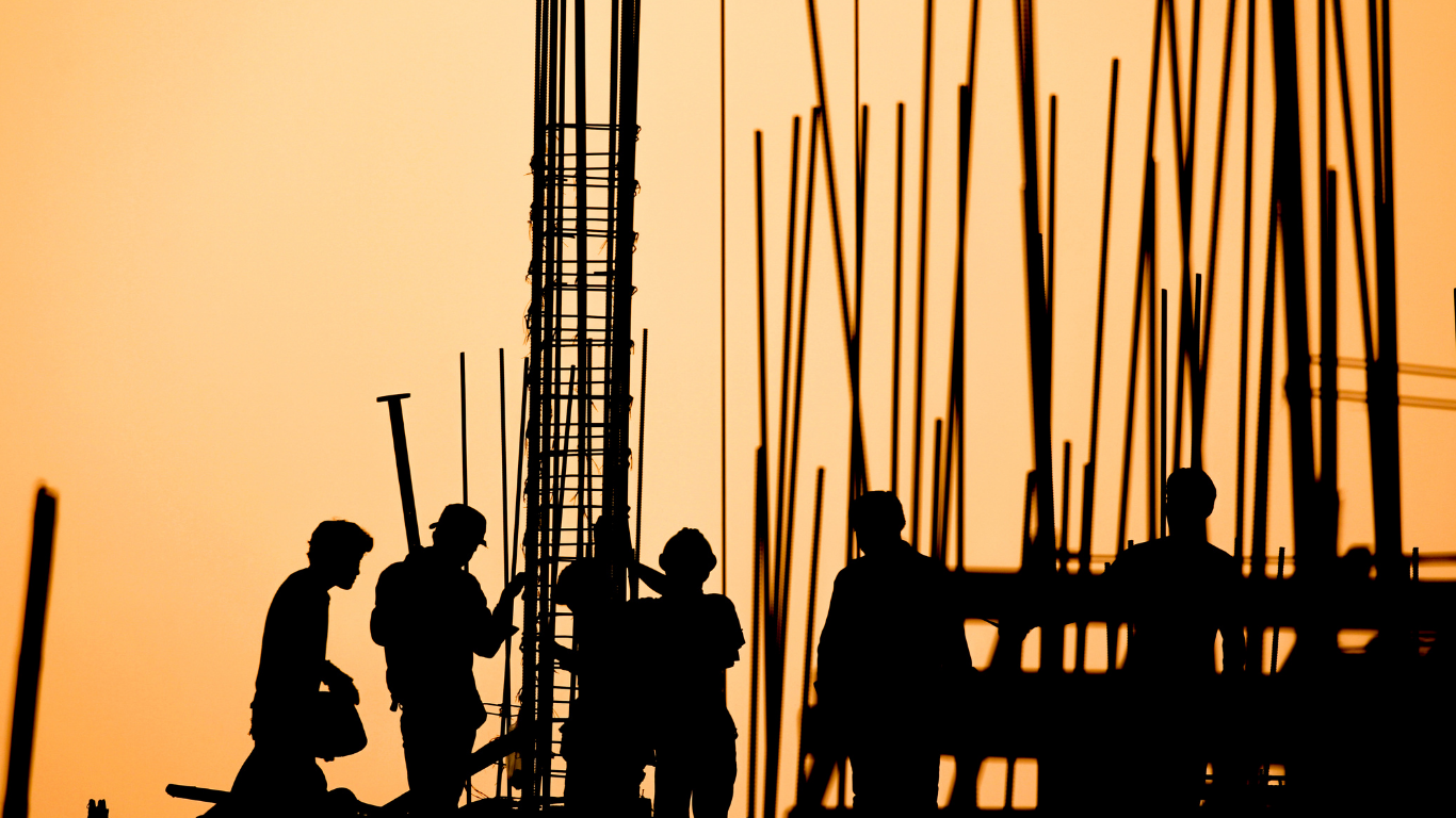 Silhouetted construction workers at a building site against an orange sky.