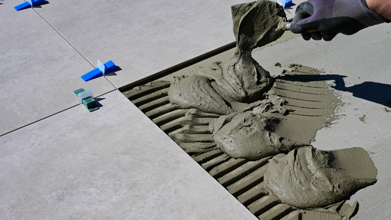 Person tiling, applying mortar with a notched trowel. Concrete surface with installed tiles and leveling clips.