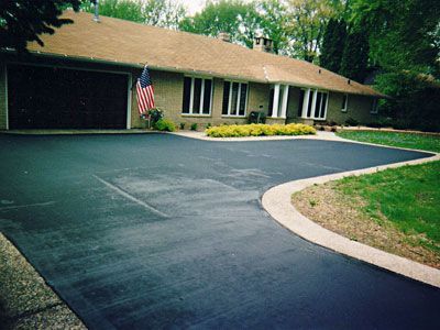 Asphalt driveway leading to a one-story house with a brown roof and a garage.