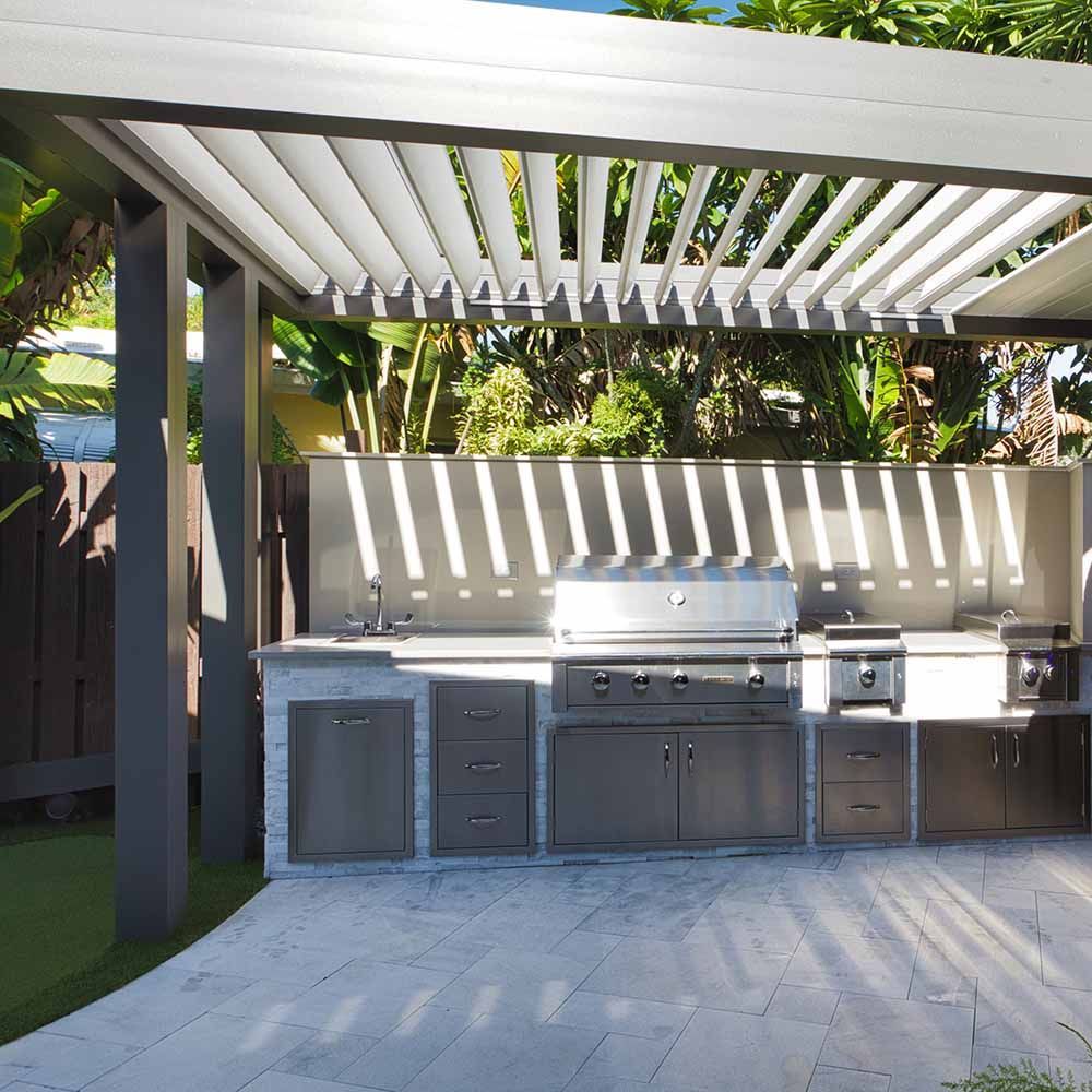 Outdoor kitchen with stainless steel appliances under a pergola, set on a stone patio.