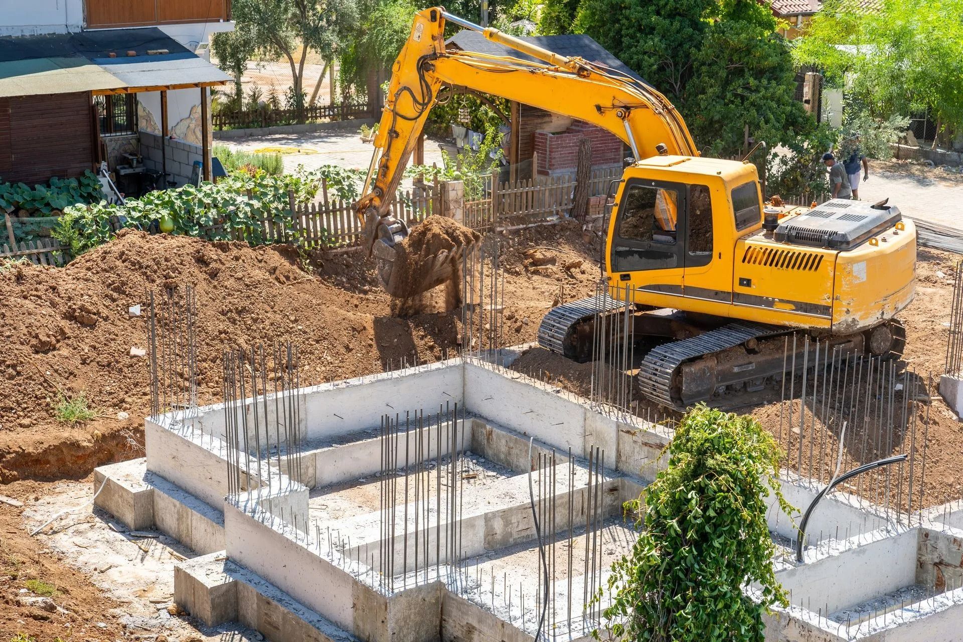 Yellow excavator digging soil at a construction site, foundation with rebar visible.