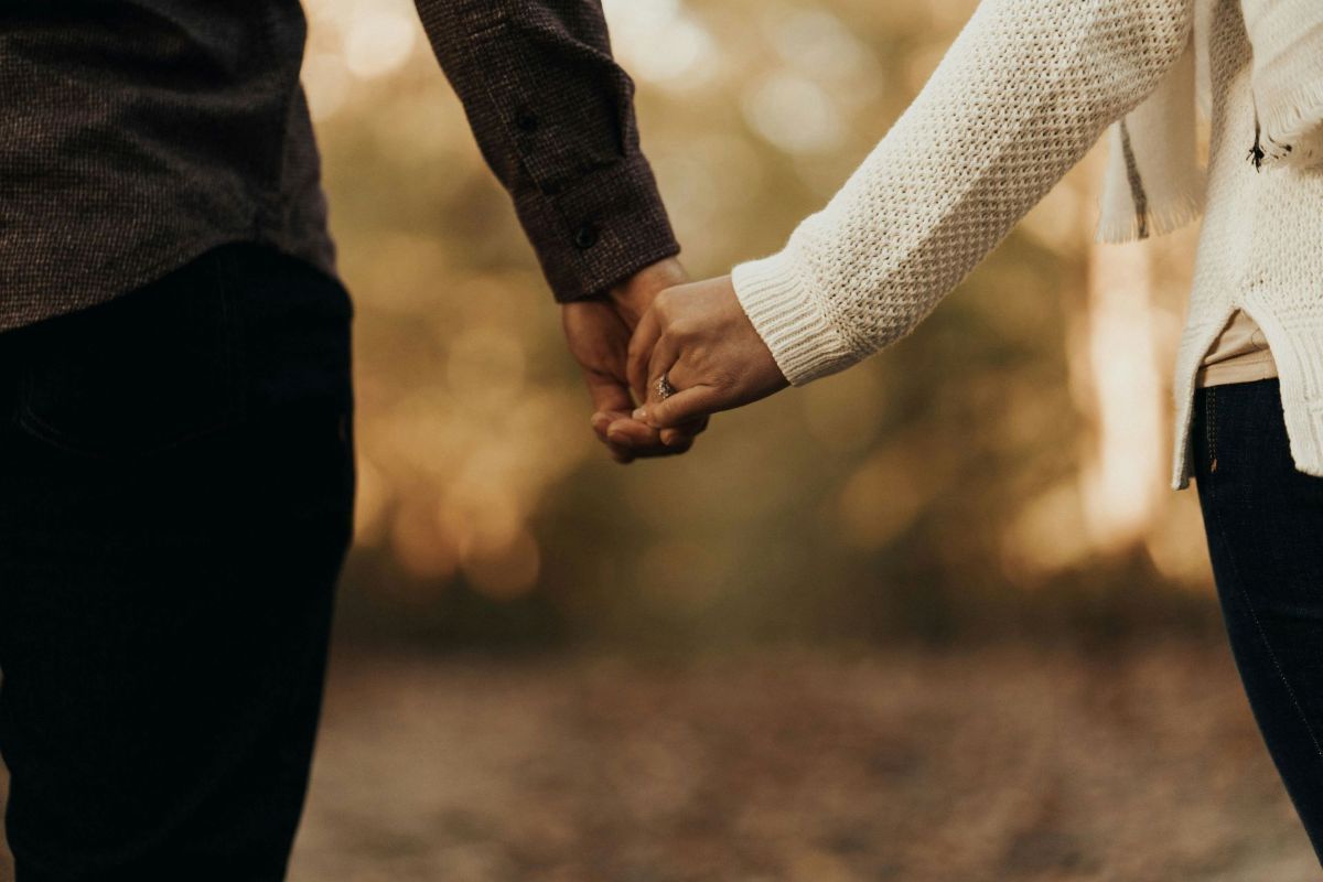 Couple holding hands outdoors. Man in brown, woman in white sweater.