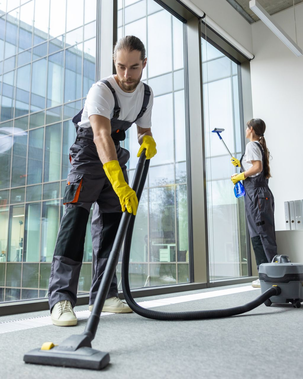 Two people cleaning office; one vacuuming carpet, the other washing windows. Both wear work overalls and yellow gloves.