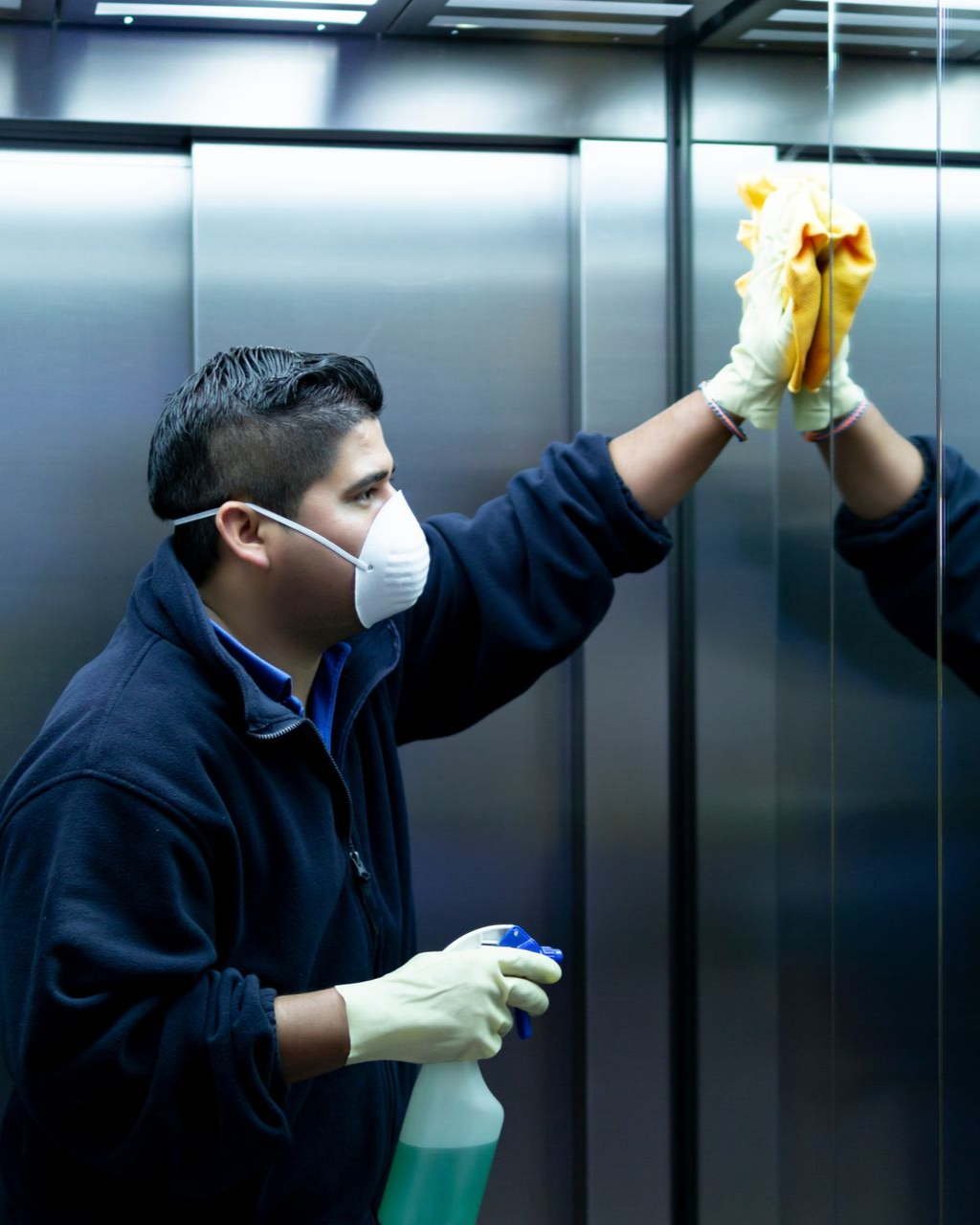Person wearing mask and gloves cleaning a mirrored elevator wall with a spray bottle and cloth.