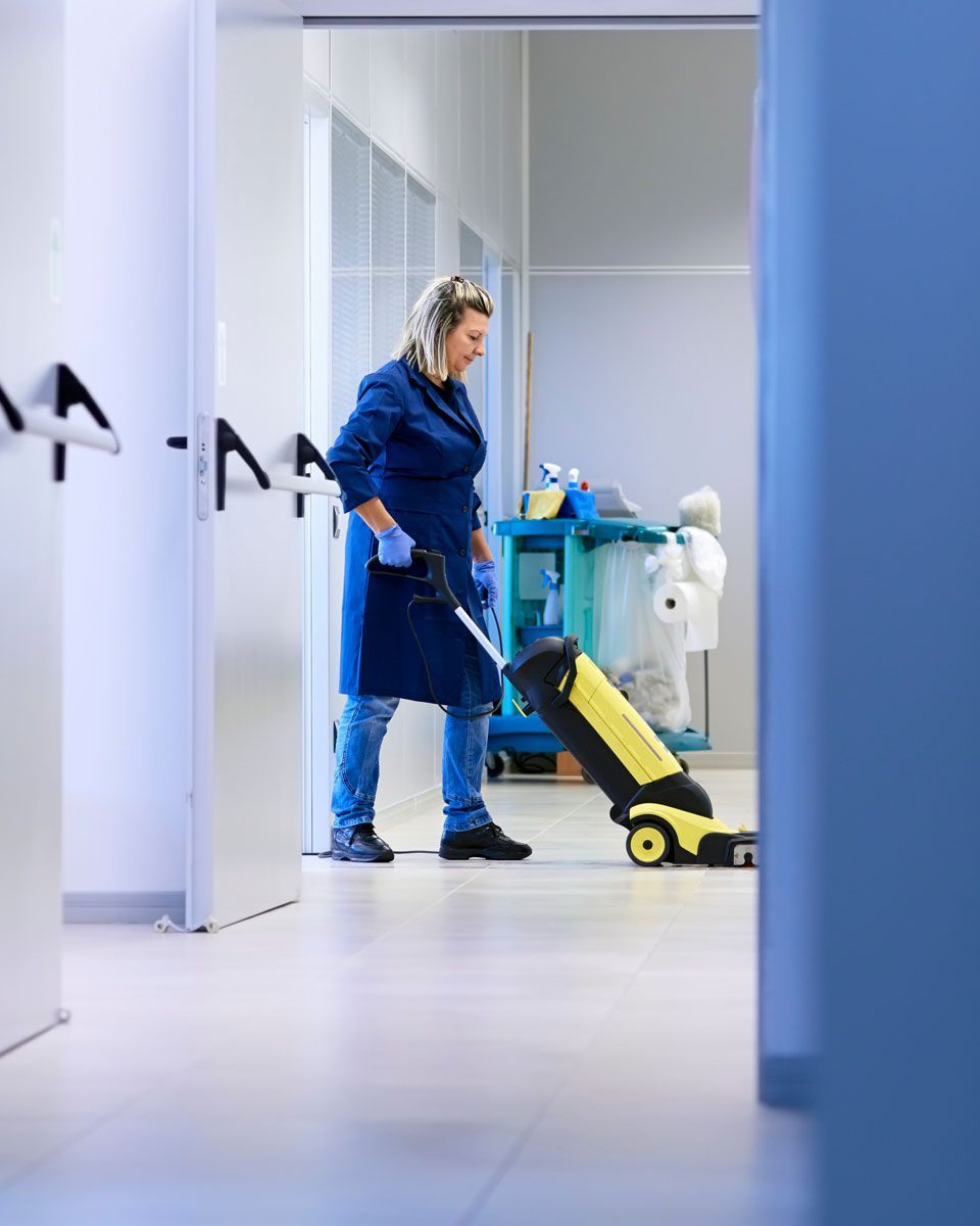 Person in blue cleaning uniform mopping a hallway with a bucket on wheels.
