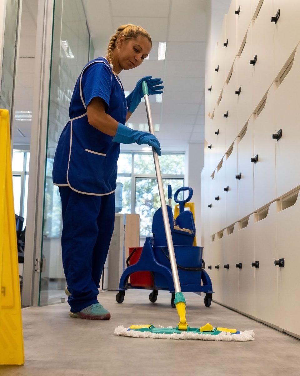 Person in blue cleaning uniform mopping a hallway with a bucket on wheels.