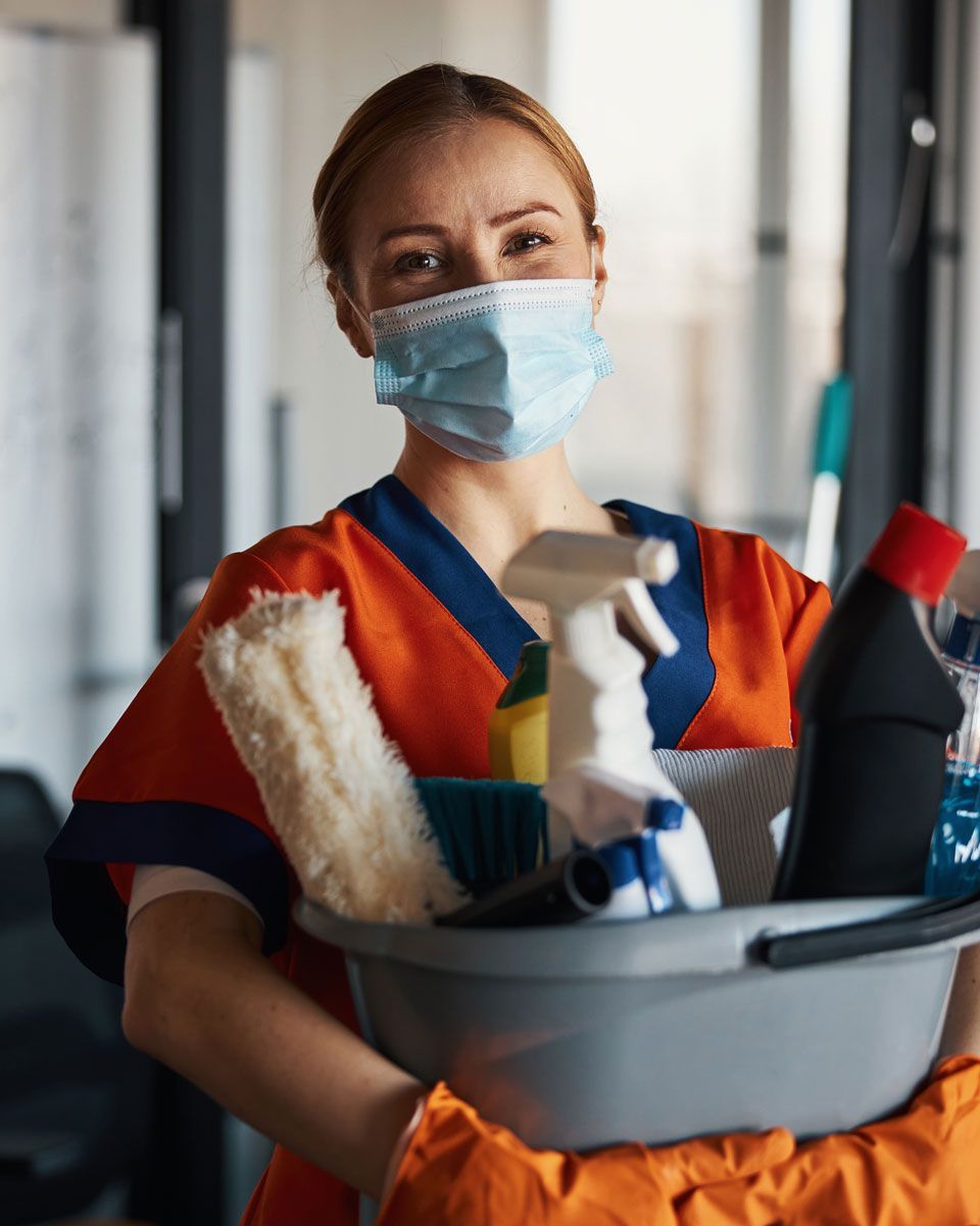 Person wearing a mask, holding cleaning supplies in a bucket. Smiling in orange uniform.