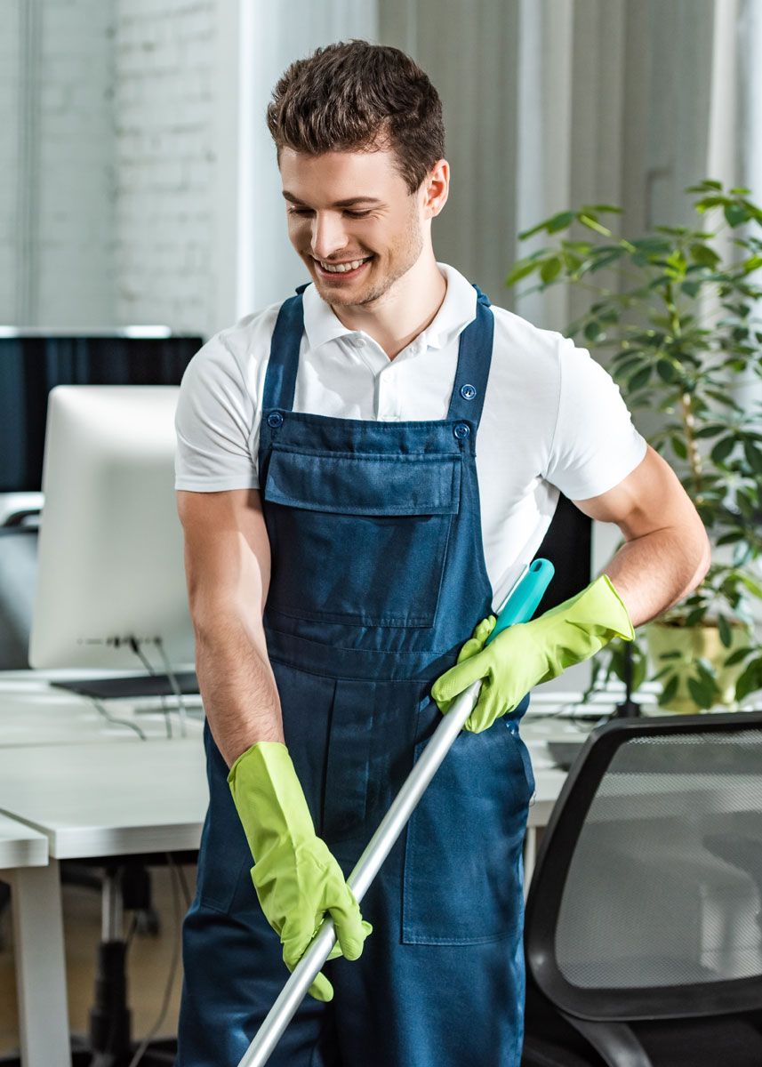 Man in overalls, wearing gloves, mopping a modern office, smiling.