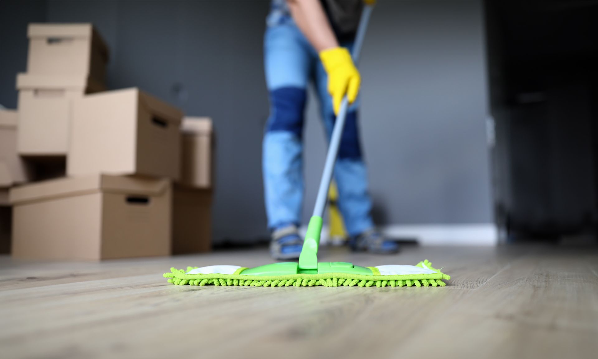 Person mopping a floor with a green mop beside stacked cardboard boxes