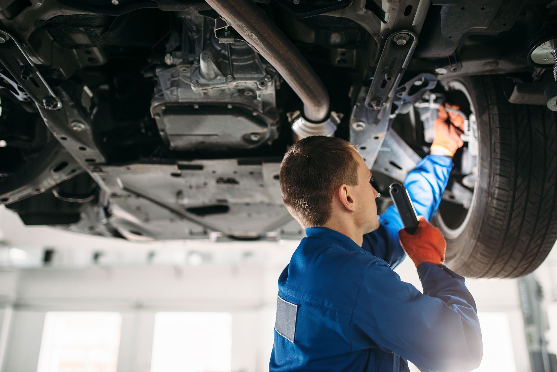Mechanic in blue overalls examines car's undercarriage, holding a flashlight in a repair shop.