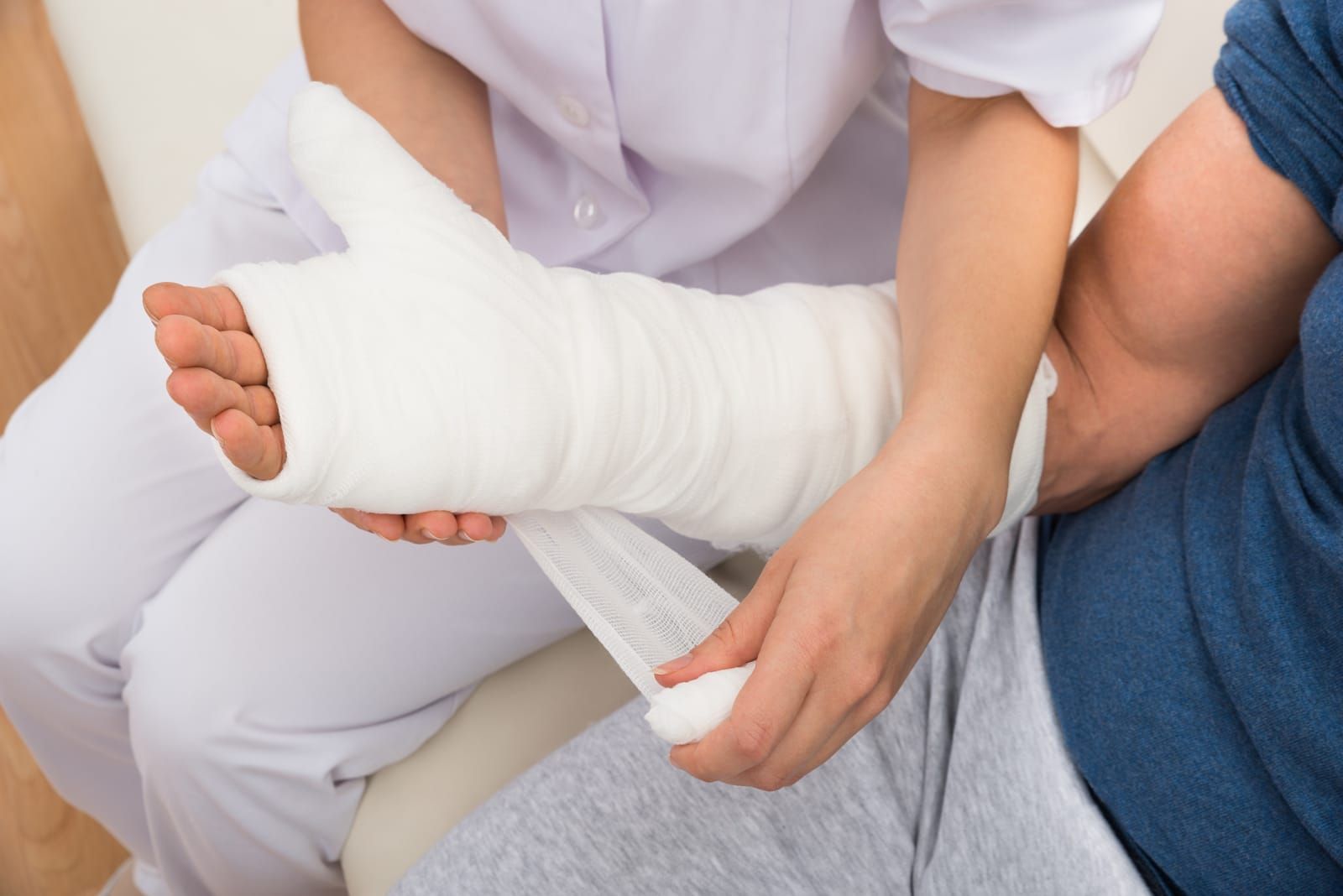 A medical professional in white scrubs wraps a bandage around a patient's arm, which is in a white plaster cast.