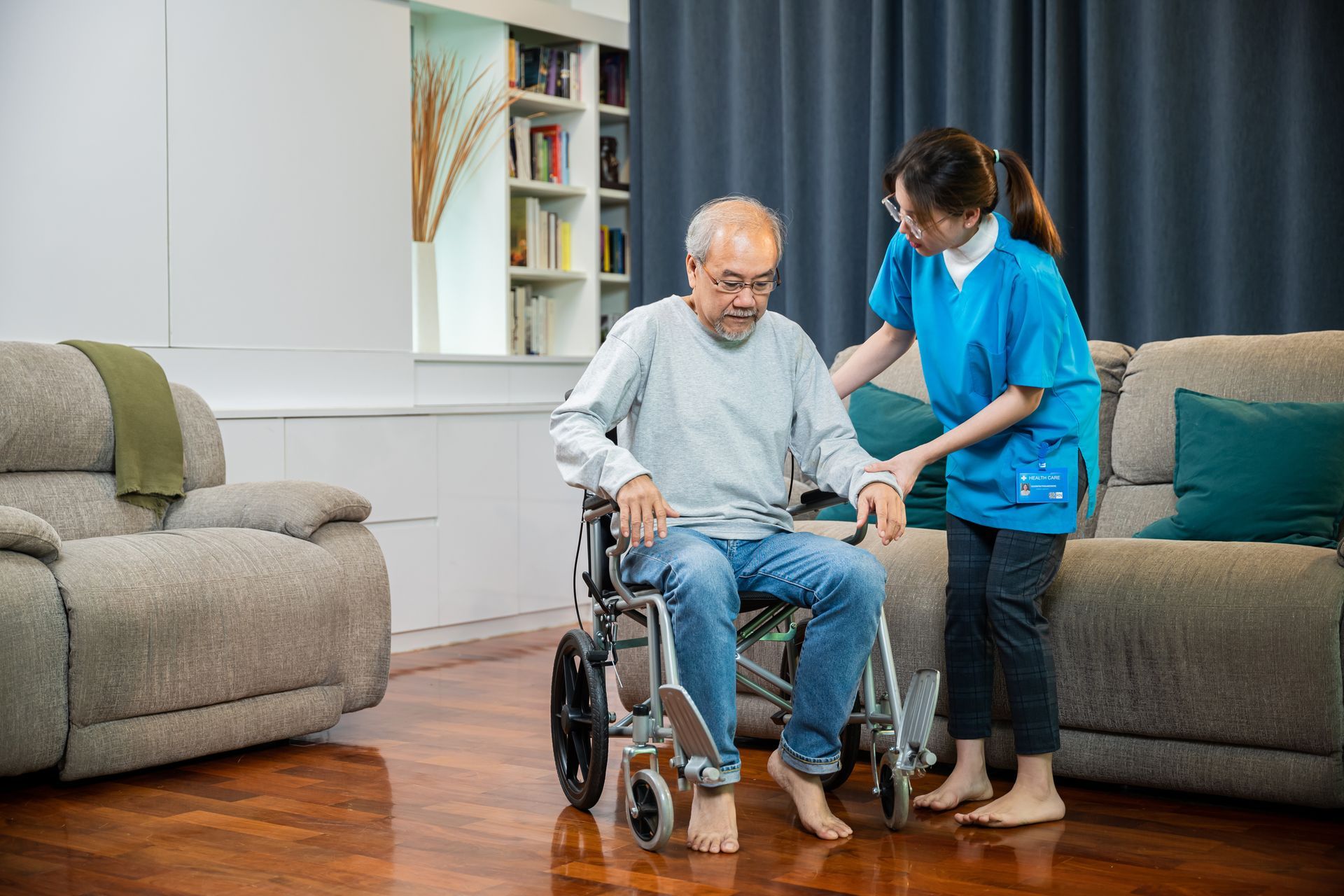 A caregiver in a blue uniform assists a man seated in a wheelchair in a living room.