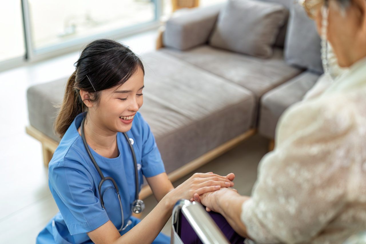 A care provider in blue scrubs kneeling and holding the hands of a person in a wheelchair in a living room.
