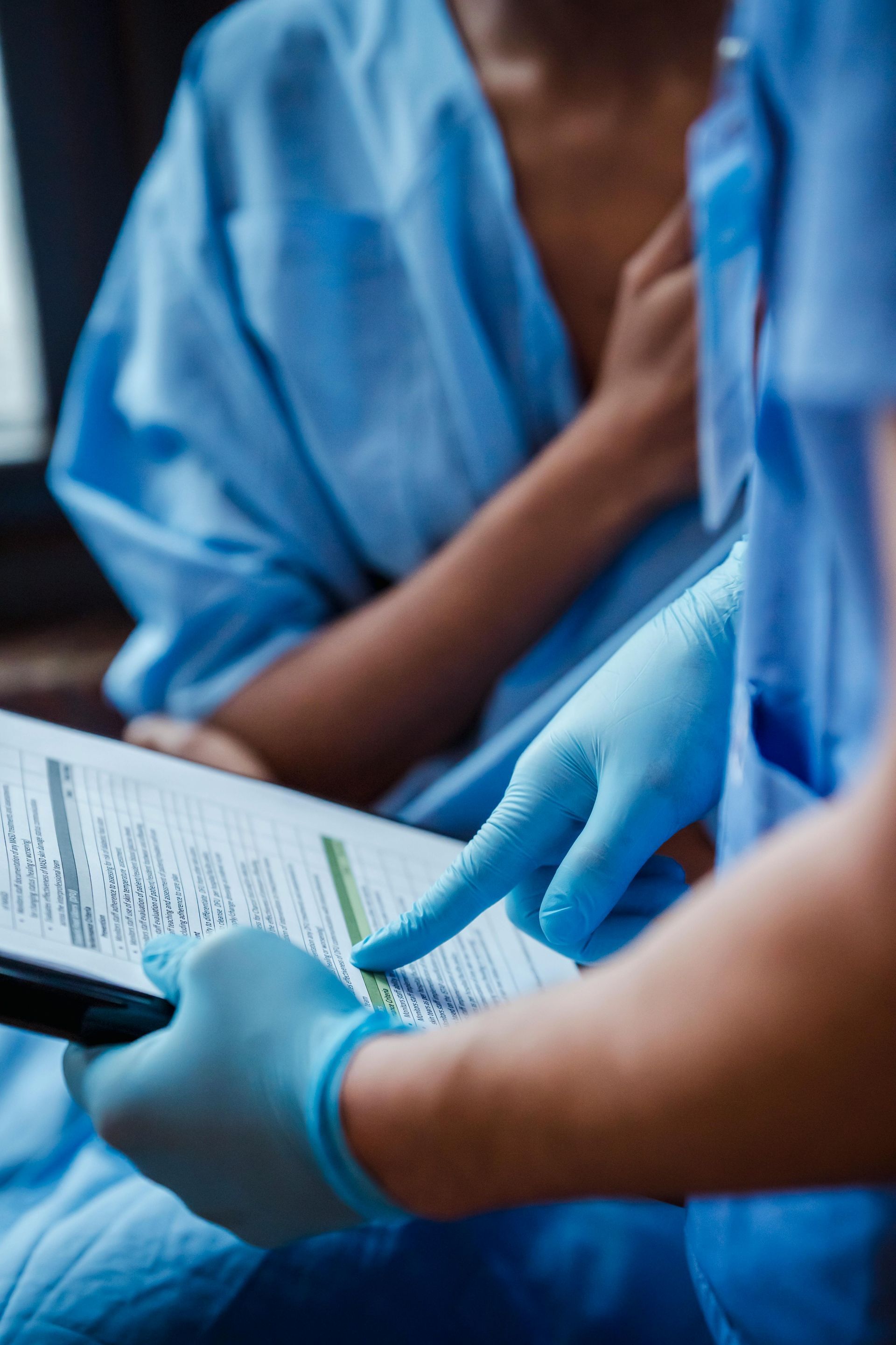 A medical professional in blue scrubs and gloves points to information on a clipboard held for a patient in a gown.