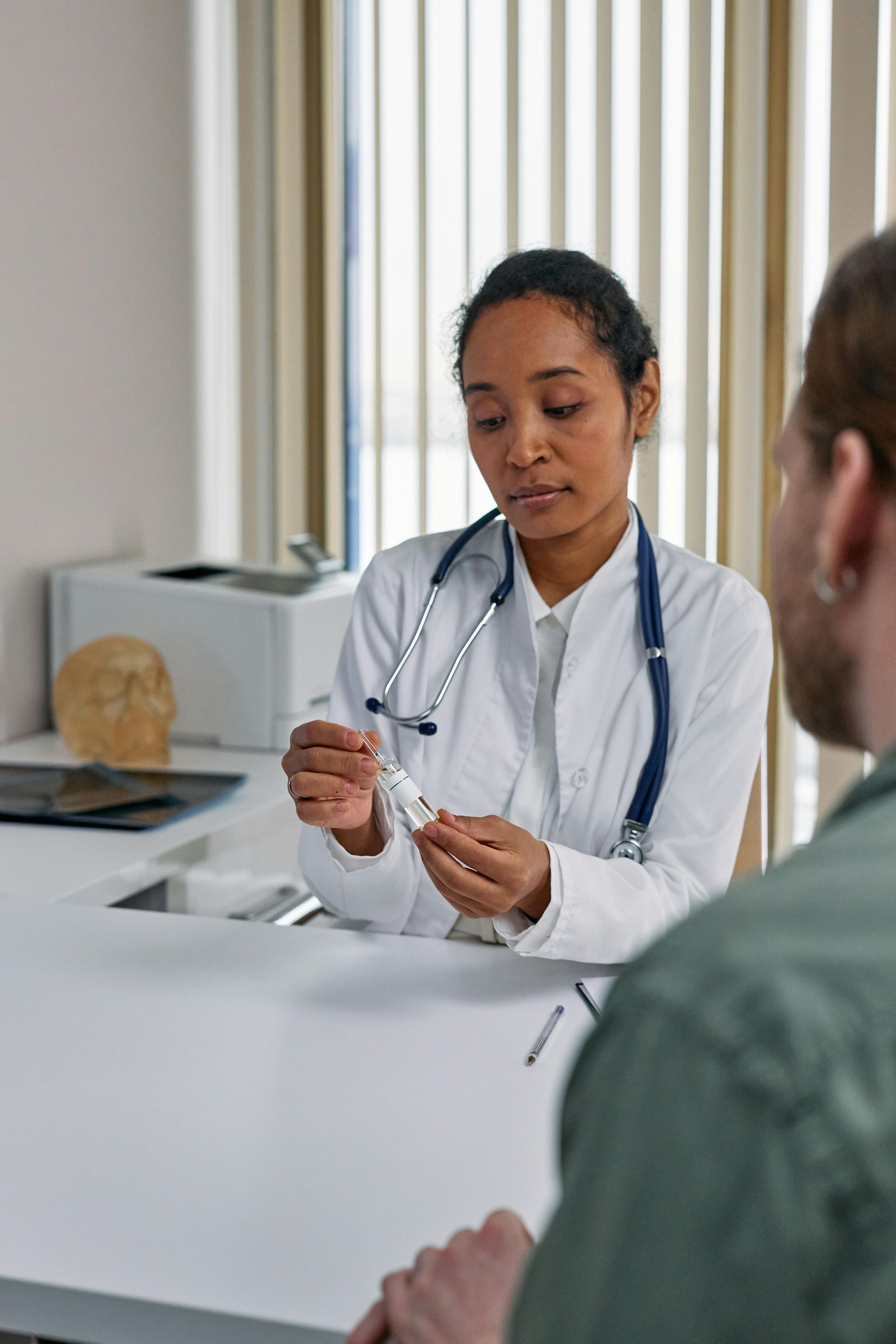 A clinician in a white lab coat with a stethoscope holds a small vial while talking to a patient in a medical office.