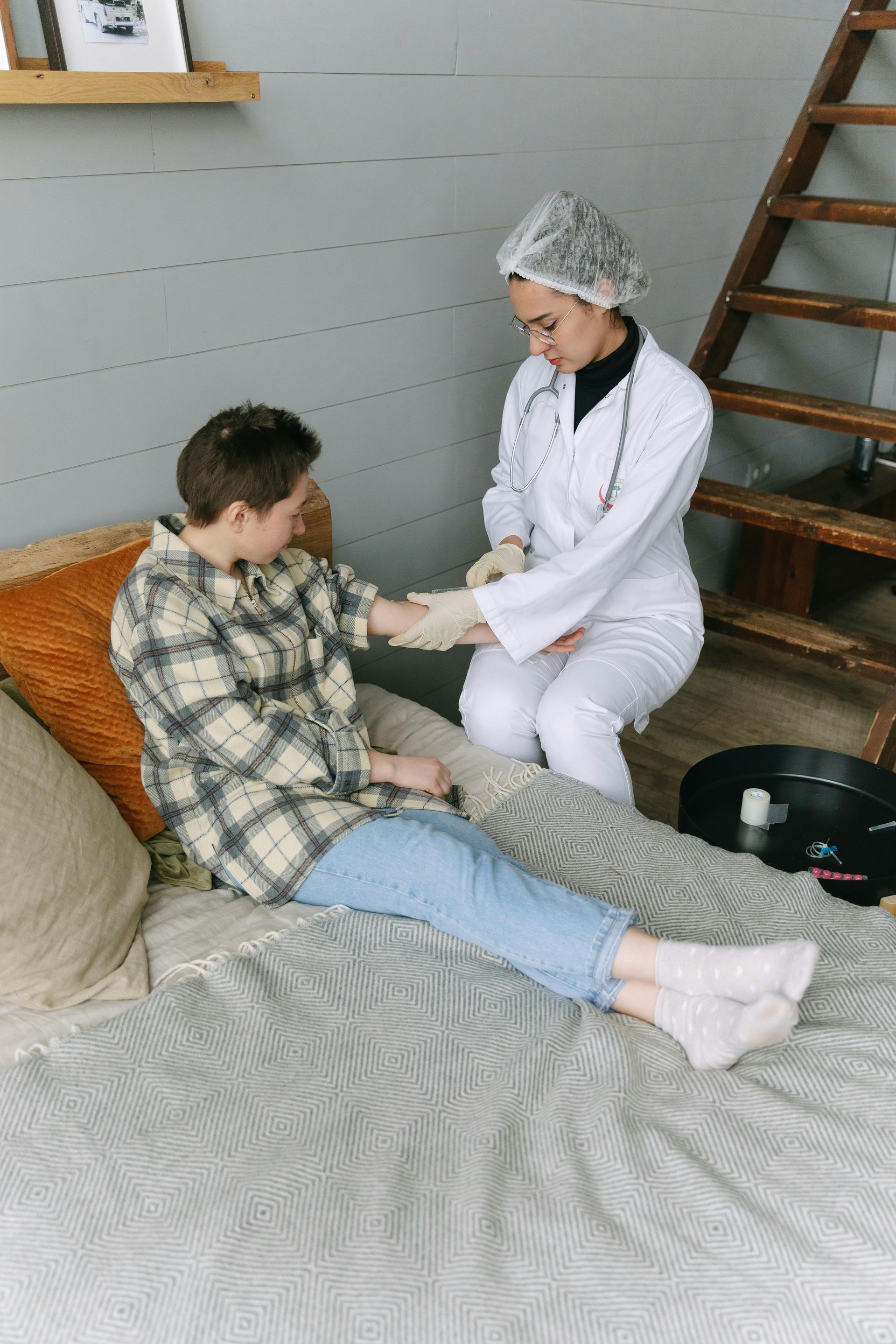 A healthcare professional in a white coat and scrub cap bandages the arm of a person sitting on a bed in a room.