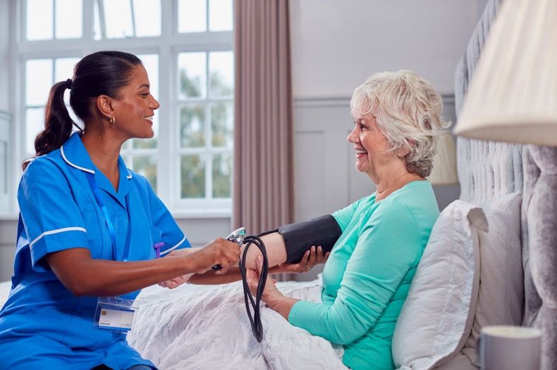 A nurse in a blue uniform takes a blood pressure reading of an elderly patient seated on a bed.