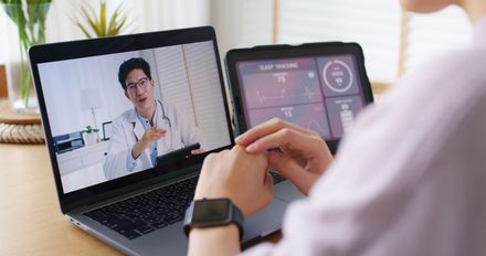 A person consults with a doctor via a laptop screen, with a digital health monitoring app open on a tablet.