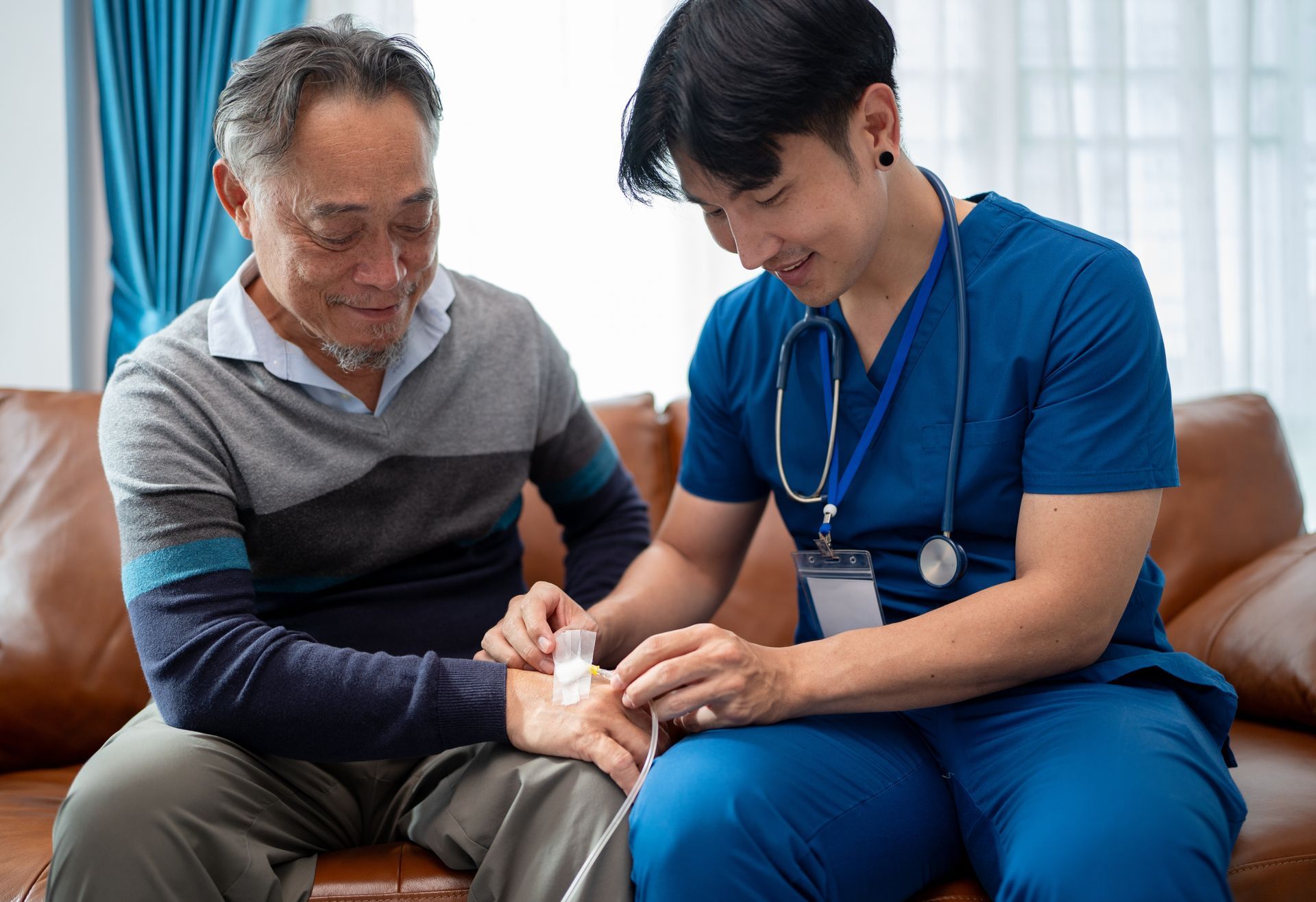 A medical professional in blue scrubs applies a bandage to a patient's hand while sitting together on a leather sofa.