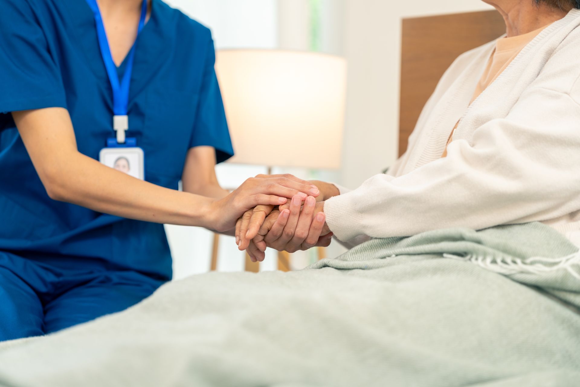 A healthcare worker in blue scrubs holds the hand of a person resting in a bed with a light-colored blanket.