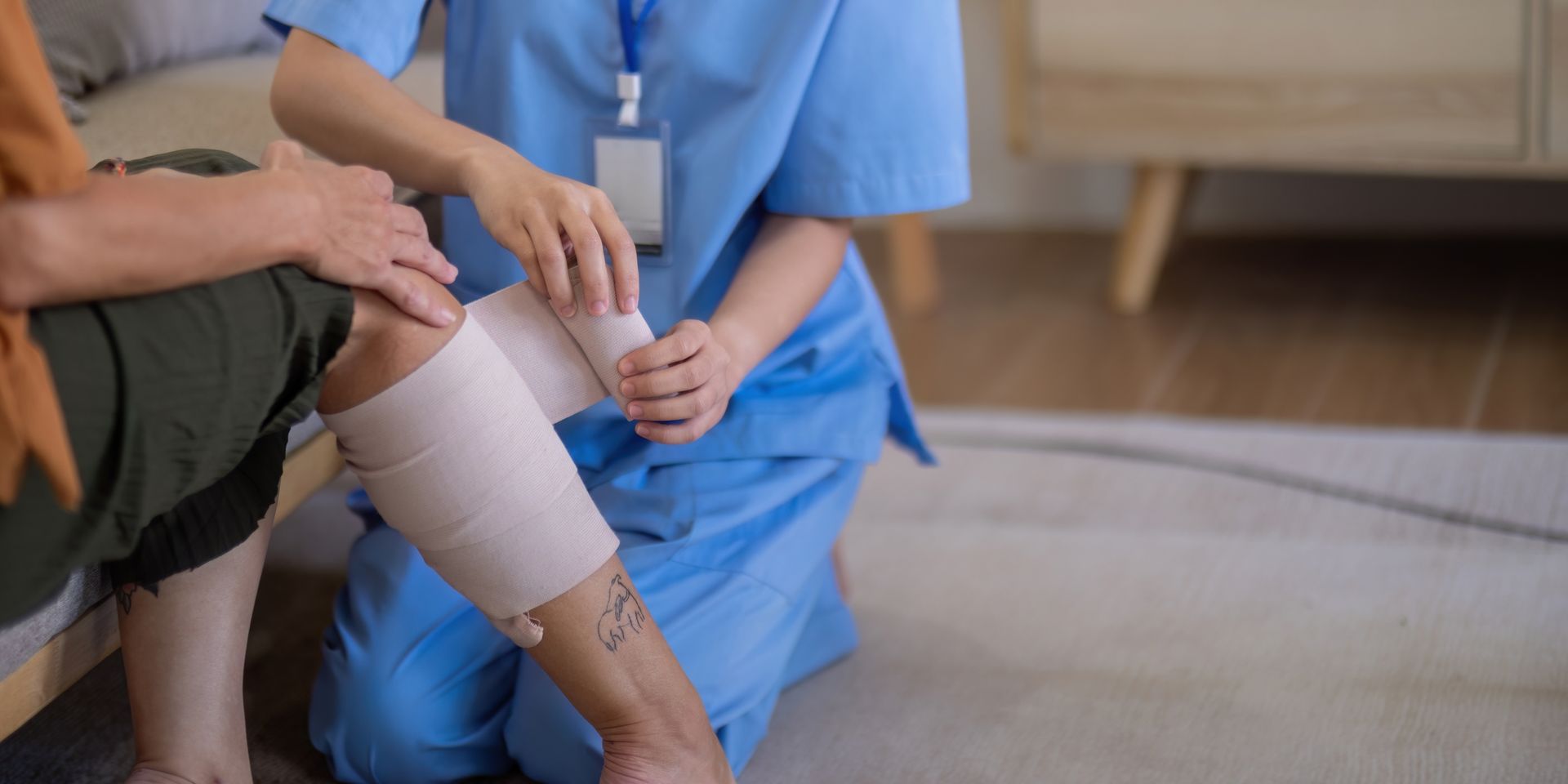 A healthcare worker in blue scrubs applies an elastic compression bandage to a patient's lower leg.