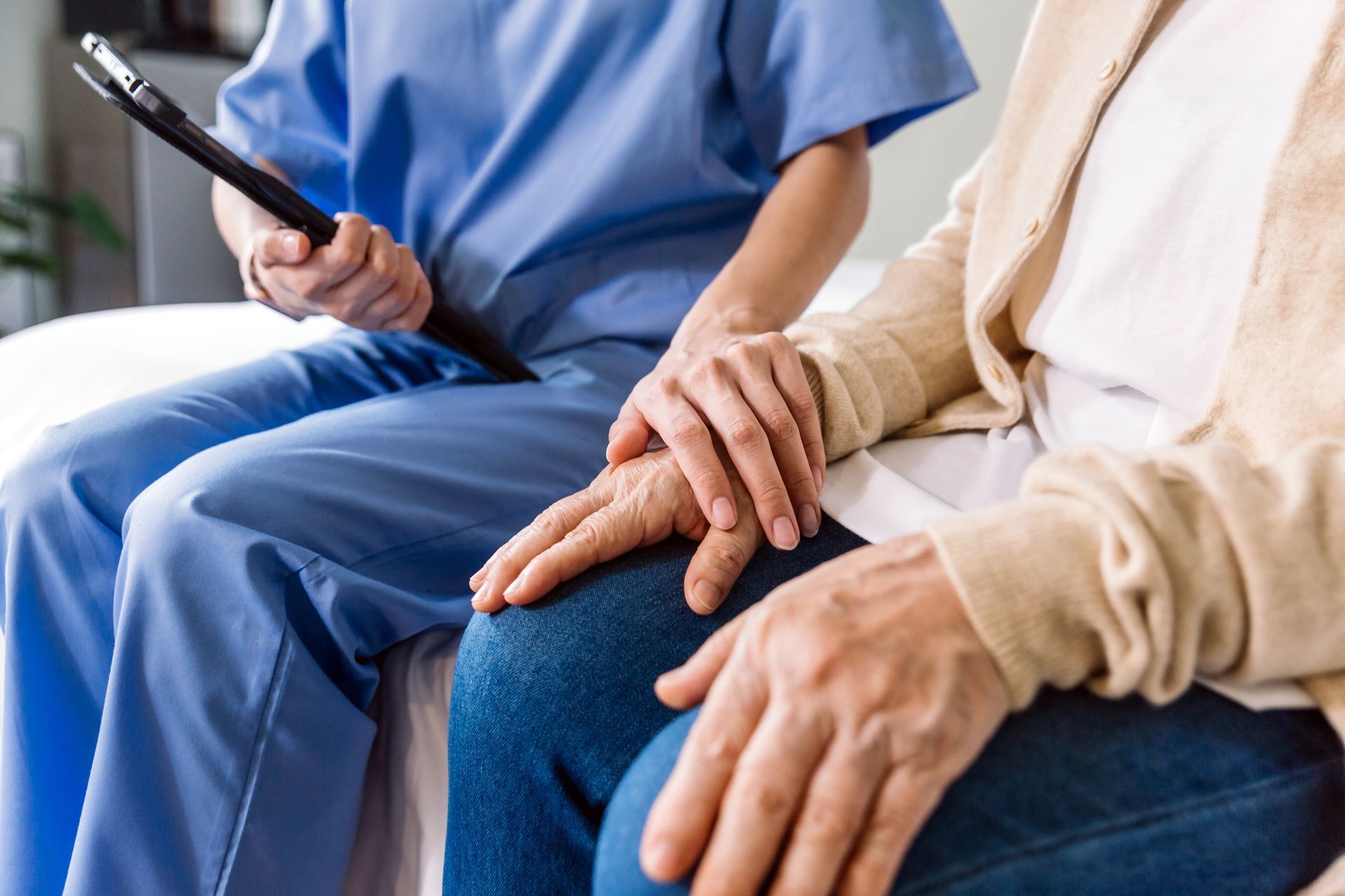 A healthcare worker in blue scrubs holds a patient's hand for comfort, with a clipboard nearby.