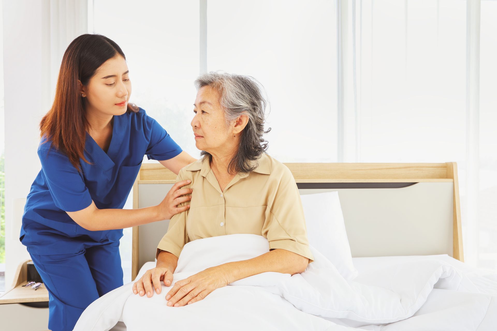 A caregiver in blue scrubs gently places a hand on the shoulder of a person sitting upright in a hospital bed.