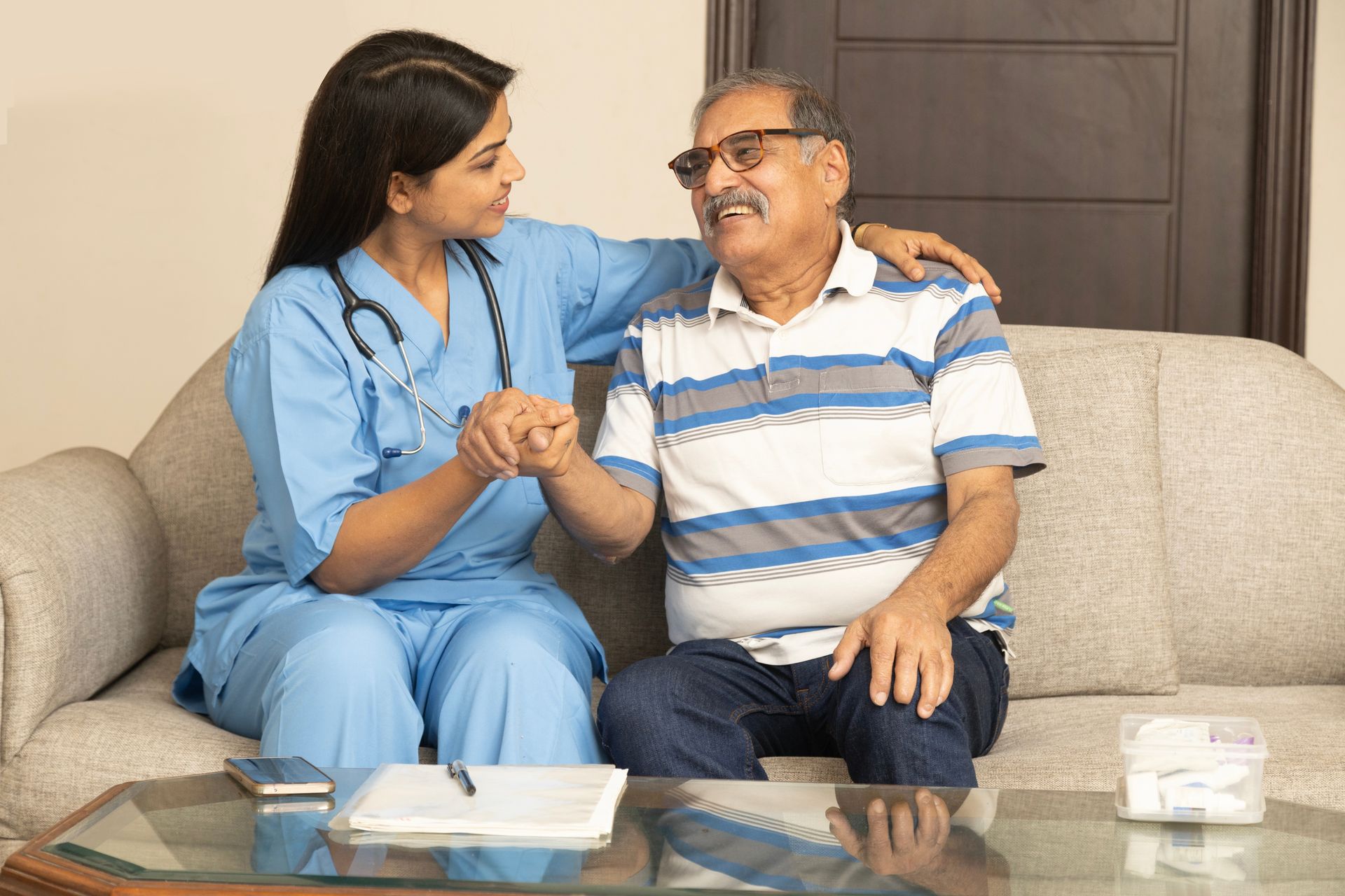 A caregiver in blue scrubs sitting on a couch and holding the hand of an older person wearing glasses.
