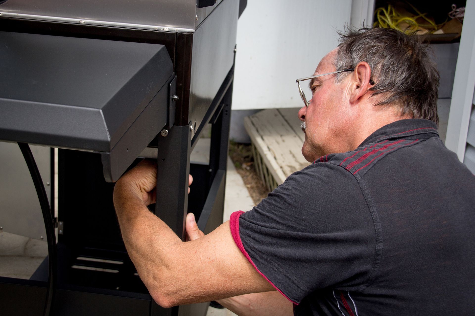 Man wearing glasses, assembling a black grill. Indoors, near white wall and outdoor steps.