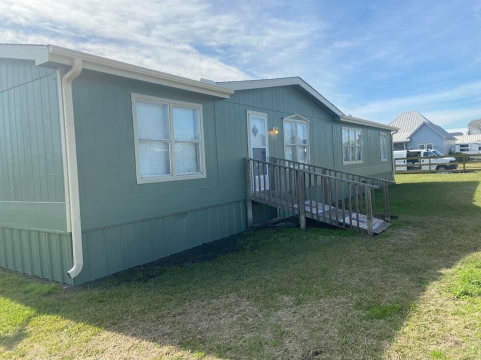 Green mobile home with a wooden ramp, on a grassy lot, under a partly cloudy sky.