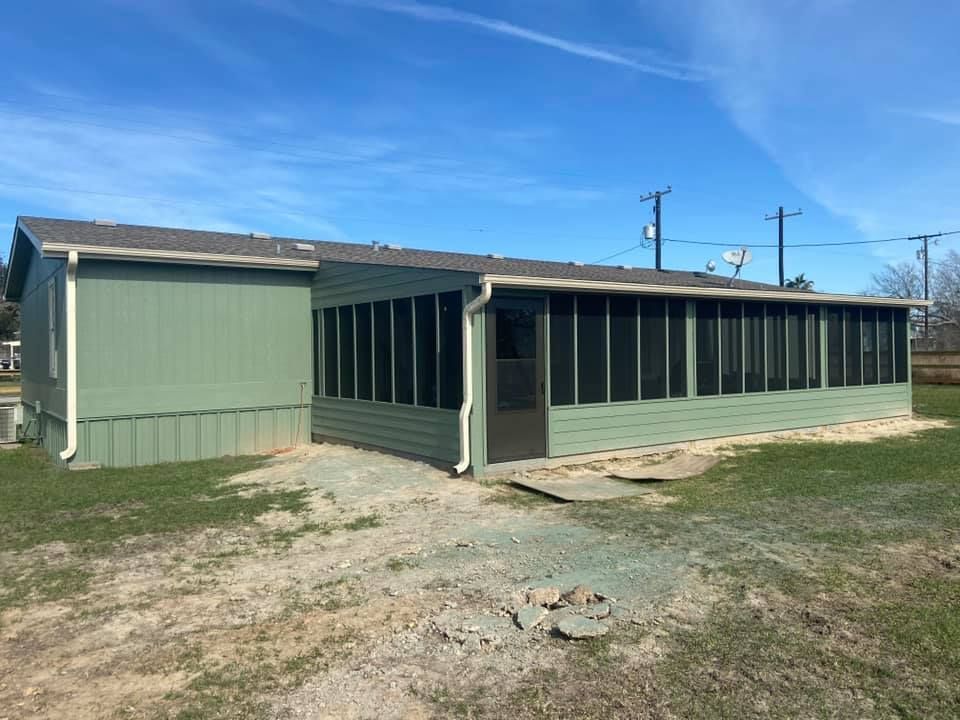Green mobile home with screened porch on a grassy lot under a blue sky.