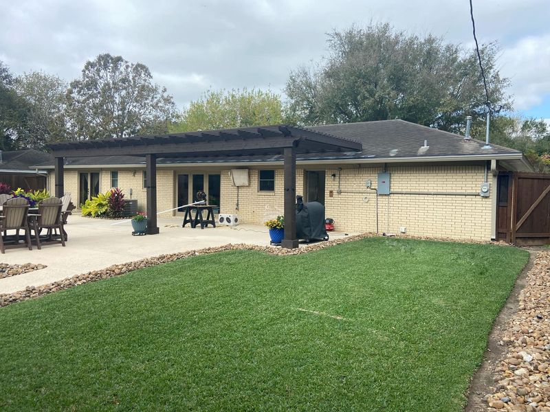 Backyard with a patio, pergola, lawn, and house. Brown pergola, beige house, green lawn, cloudy sky.