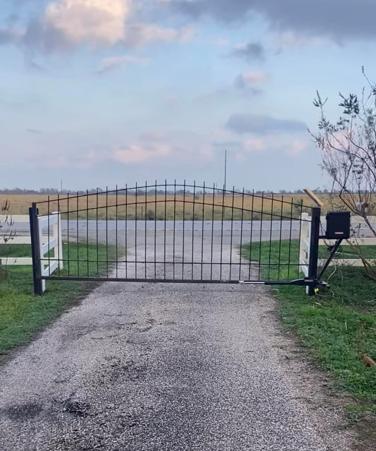 Black metal gate on a gravel driveway, with white posts and an automated opener, in a rural setting.