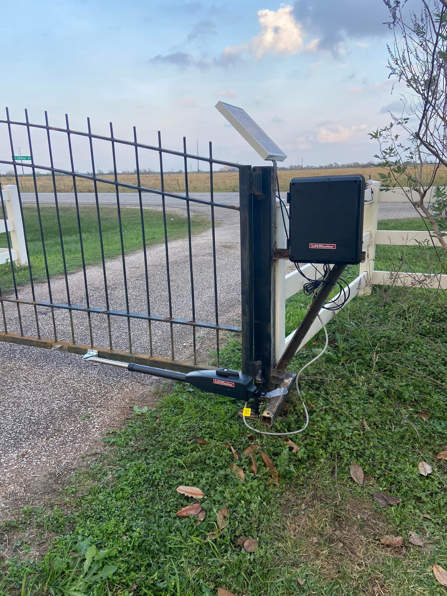 Black automated gate with solar panel and control box, in a grassy area with a fence and road.
