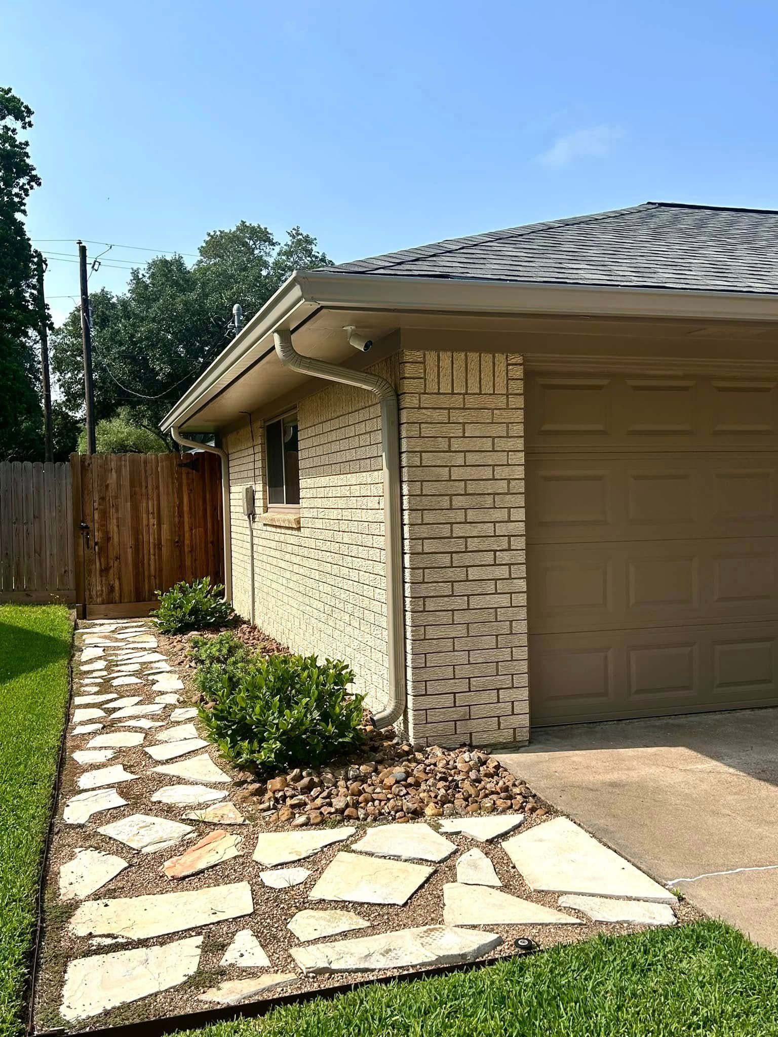 Stone path leads to a garage with a tan door and light brick exterior; green grass and bushes surround.