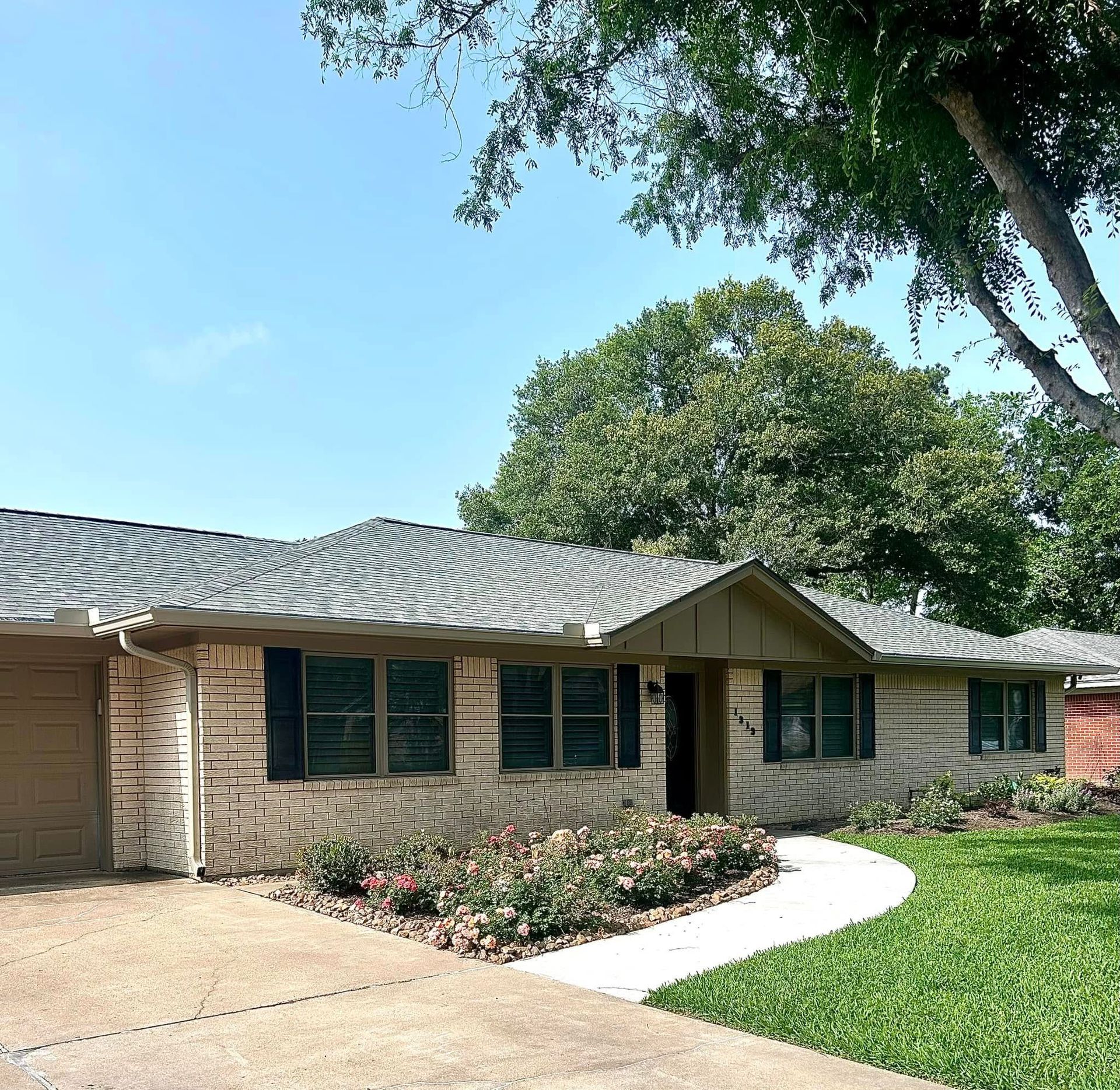 Single-story brick house with a brown garage door and black shutters. A walkway curves to the front door. Green lawn.