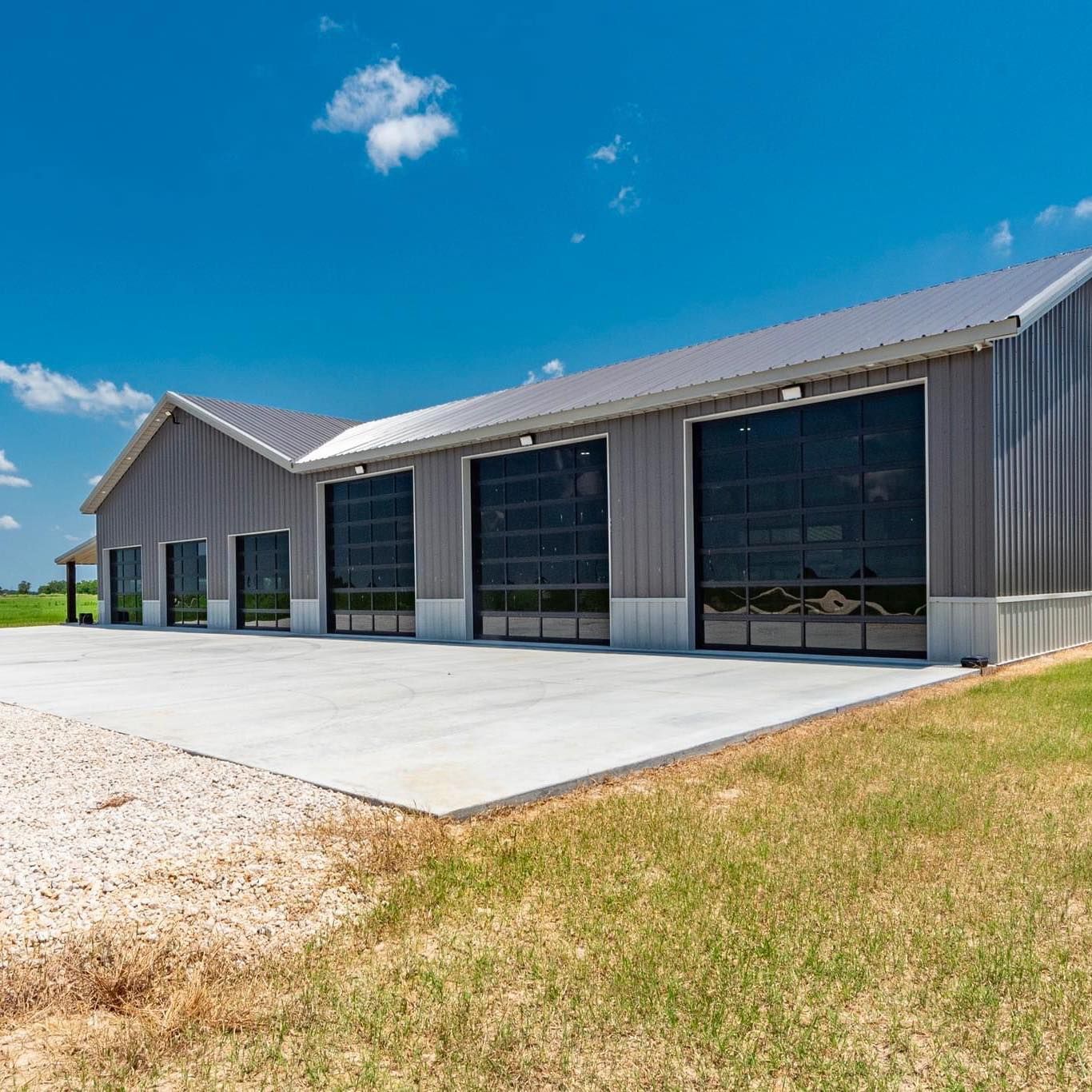 Gray metal building with large glass garage doors, on concrete, under a blue sky.