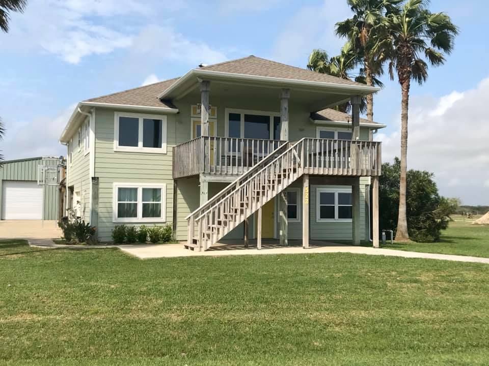 Two-story light green house with wooden deck, staircase, and palm trees in a grassy yard under a blue sky.
