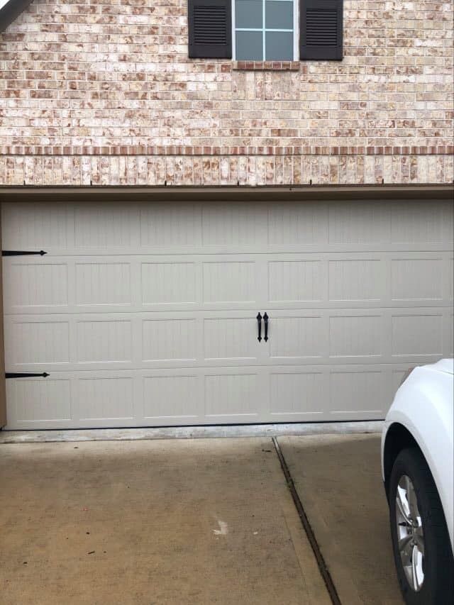 Beige garage door with black hardware, brick facade, and car parked partially in driveway.
