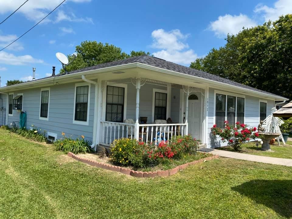 Light blue house with white porch, flower beds, and a green lawn under a partly cloudy sky.
