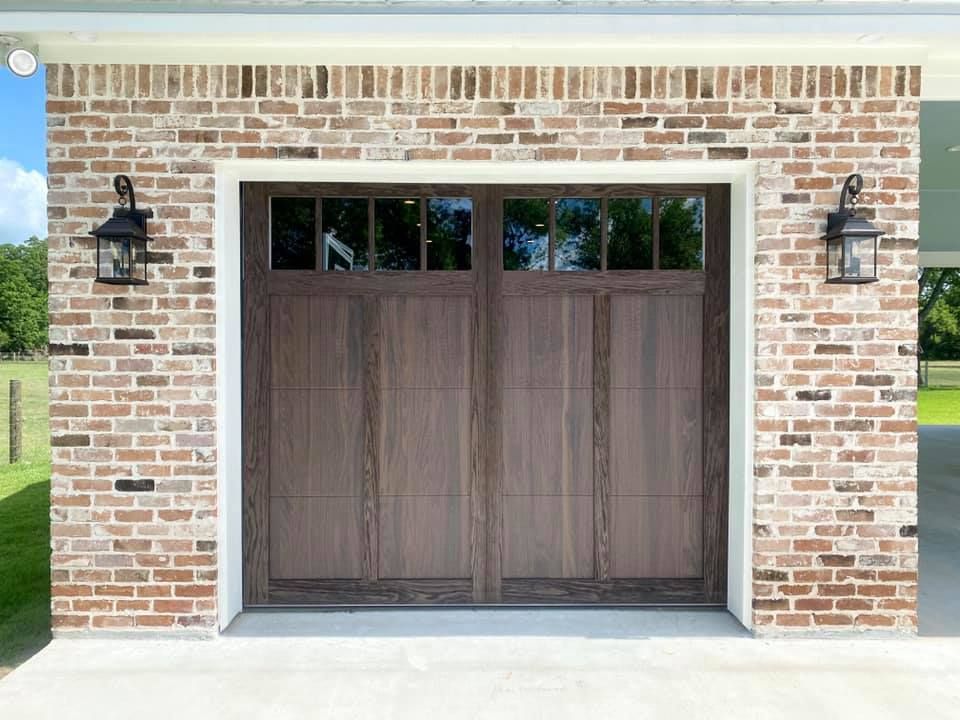 Brown garage door with windows, flanked by brick, and lanterns on a white trimmed doorway.