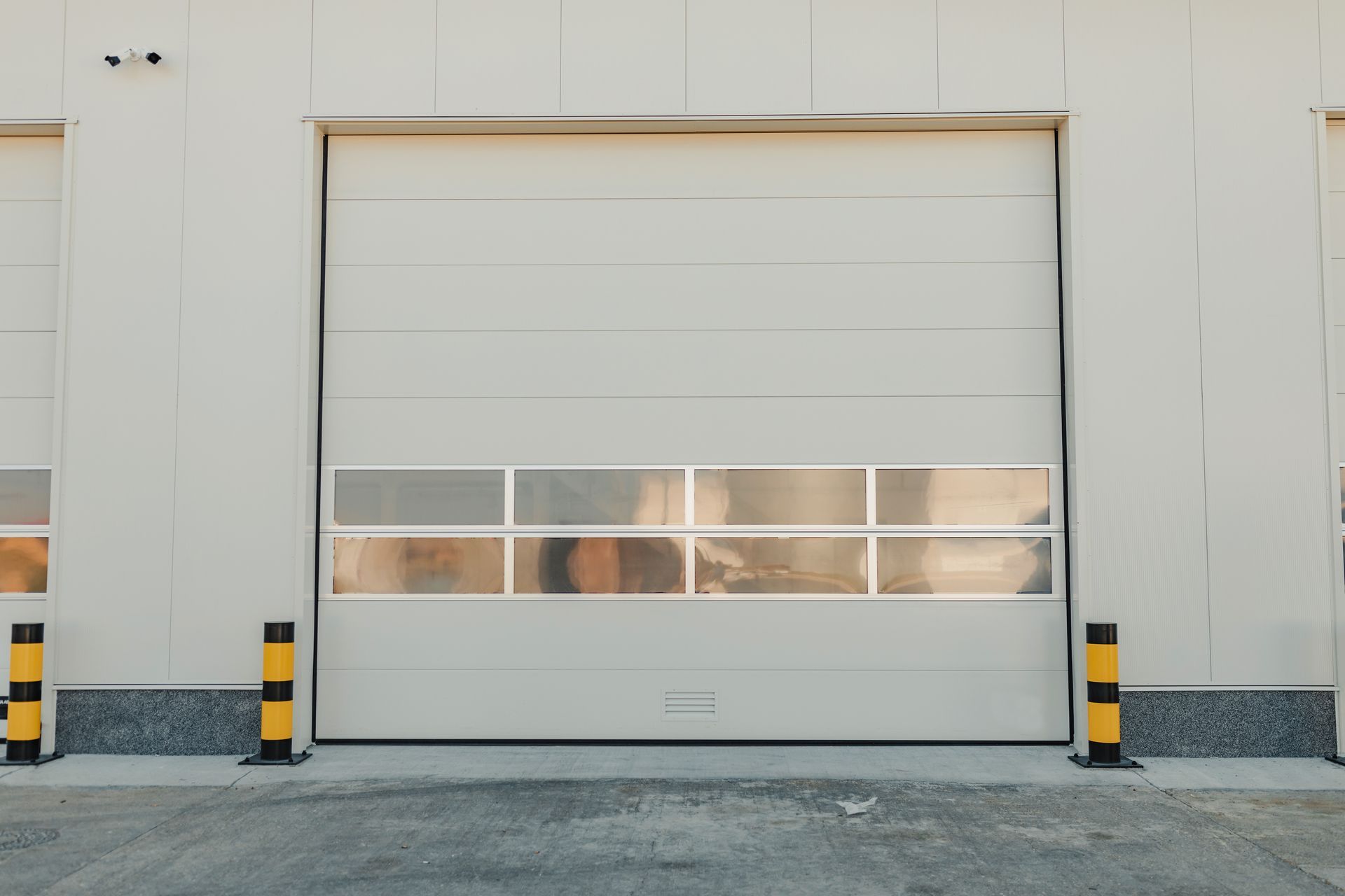 Large, closed white industrial garage door with a clear panel, flanked by yellow and black bollards.