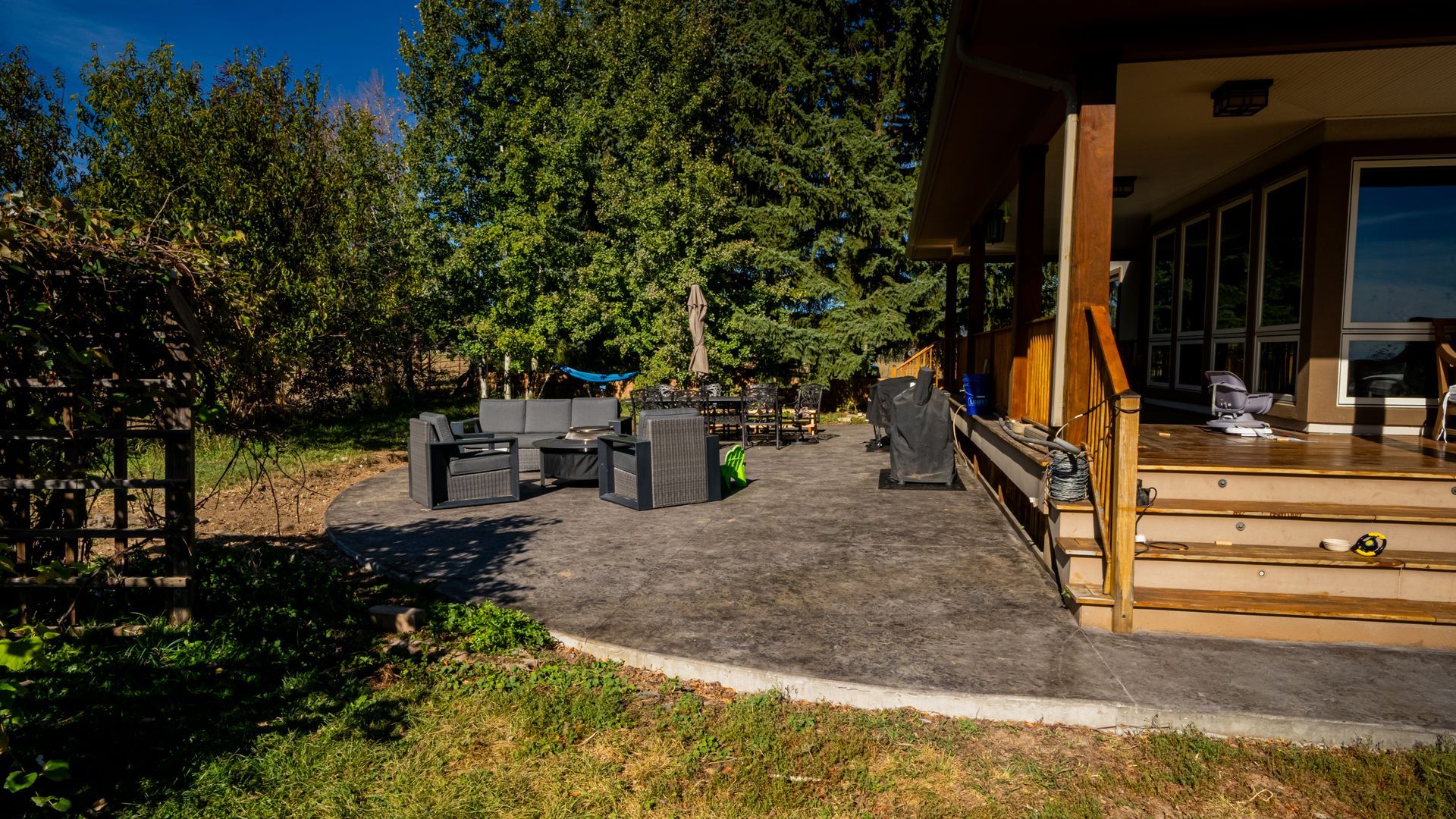 A modern outdoor patio with built-in wooden bench, cushions, and a tree, alongside a white wall with sliding glass doors.