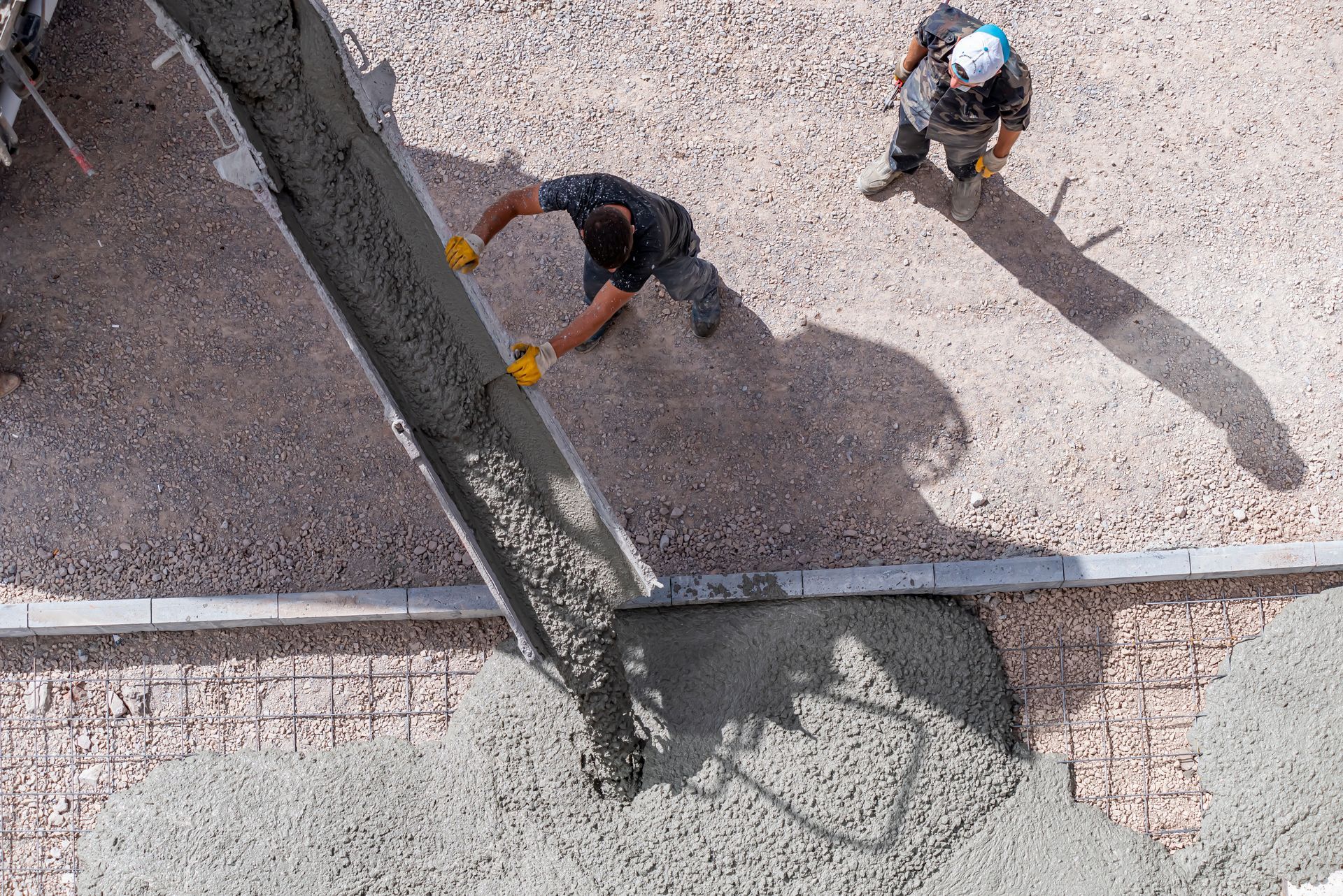 Construction workers pouring concrete on a gravel surface. One worker guides the flow, another watches.