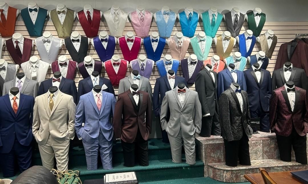 Suits displayed on mannequins in a clothing store, with colorful vests hanging on the wall behind.