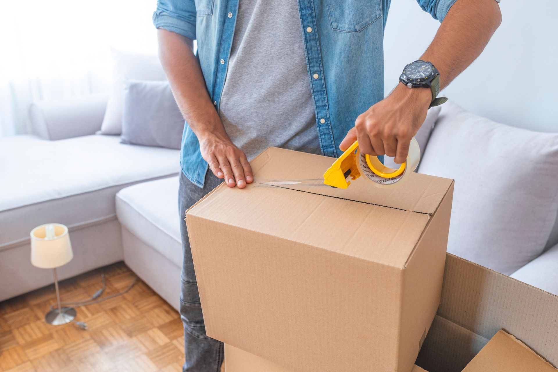Man taping cardboard moving boxes with packing tape dispenser for home relocation.