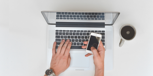 A person is typing on a laptop computer while holding a cell phone.