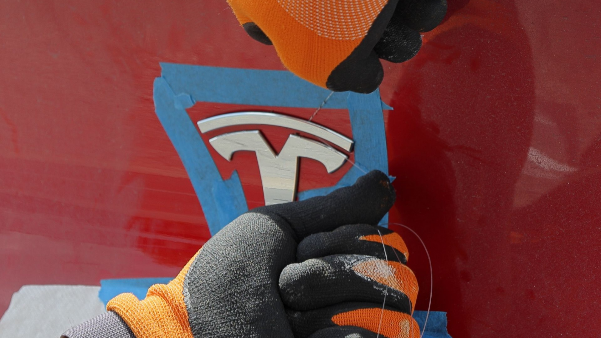 Gloved hands peeling blue tape from a red Tesla logo on a car panel